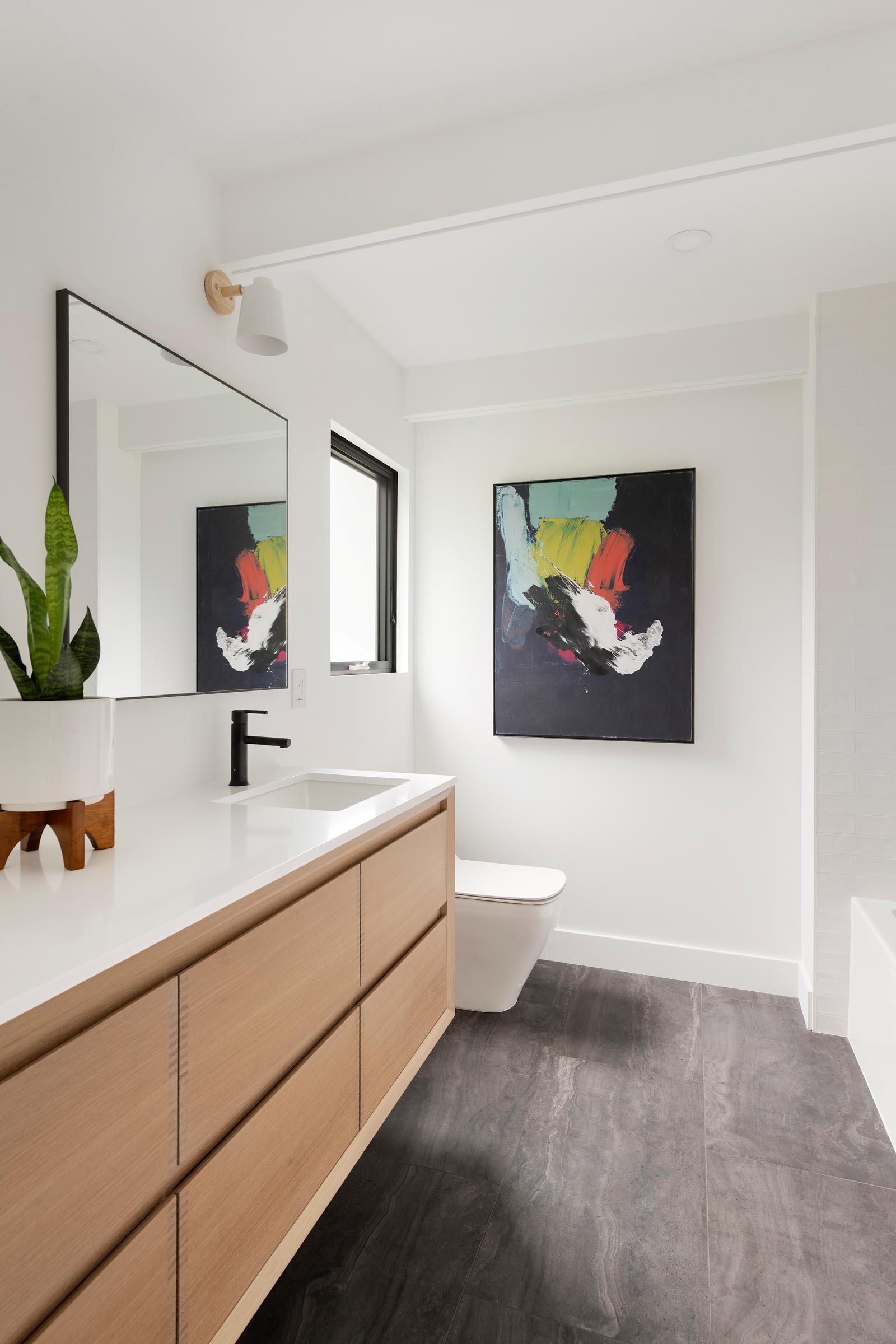 In this modern bathroom, white walls and a light wood vanity keep the space bright, and artwork adds a colorful element.