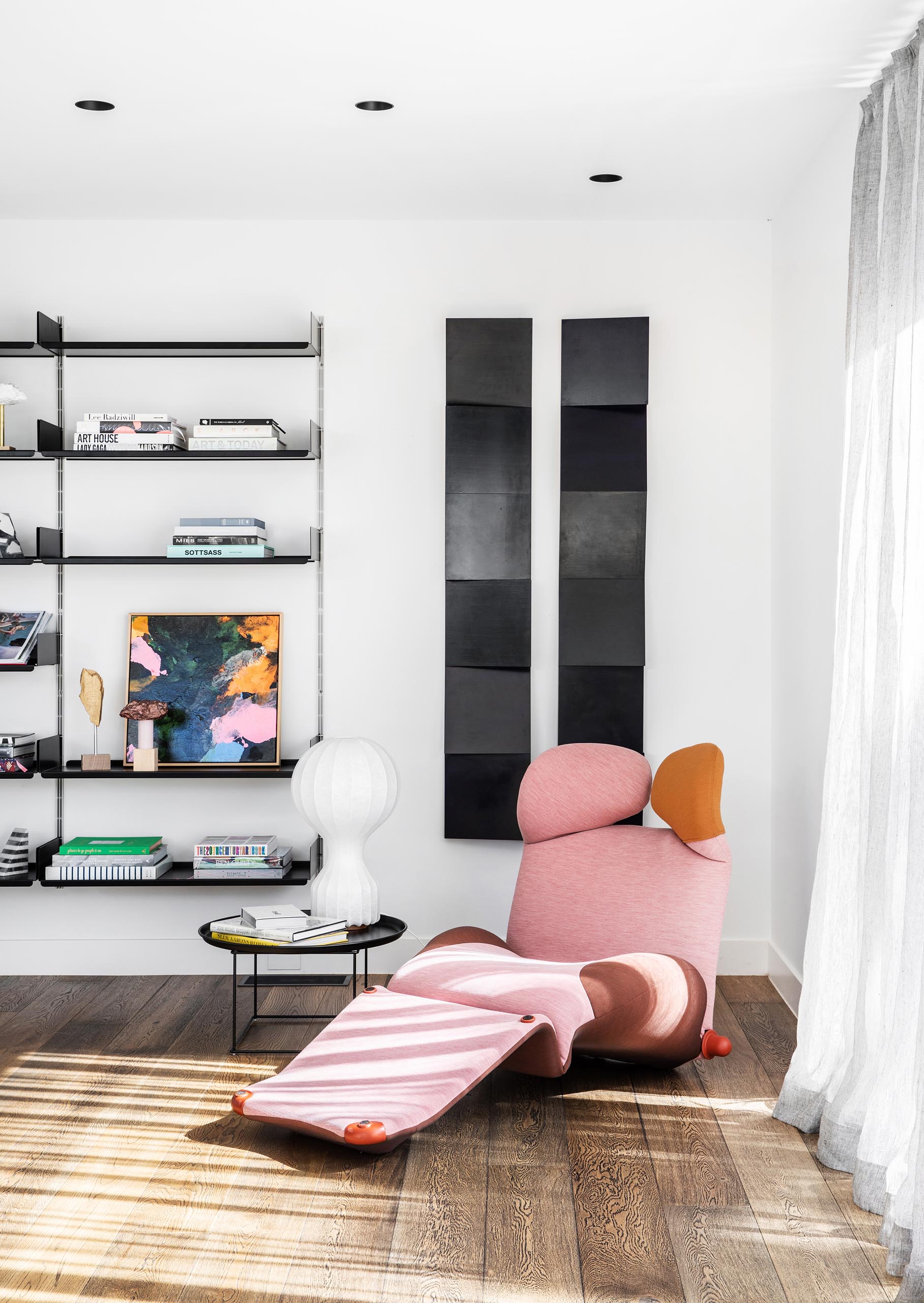 Tucked into the corner behind the living room is a small reading nook that includes a sculptural pink chaise, vertical art pieces, and a wall-mounted bookshelf.