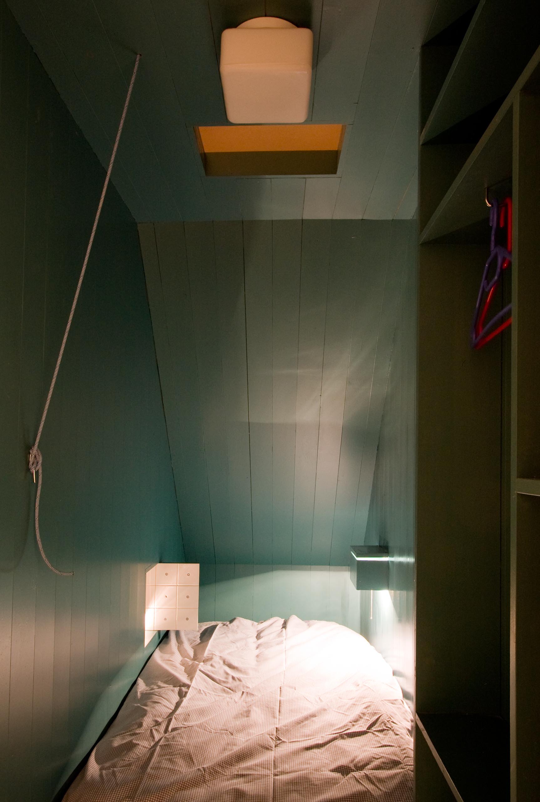 A modern turquoise blue bedroom with a small window that looks out to the living room.