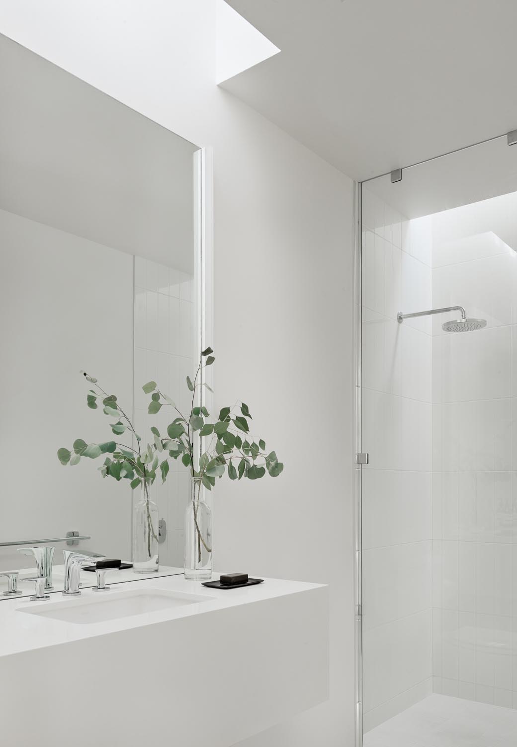 A modern white bathroom with a skylight that highlights the vanity, and a floor-to-ceiling glass screen separates the shower.