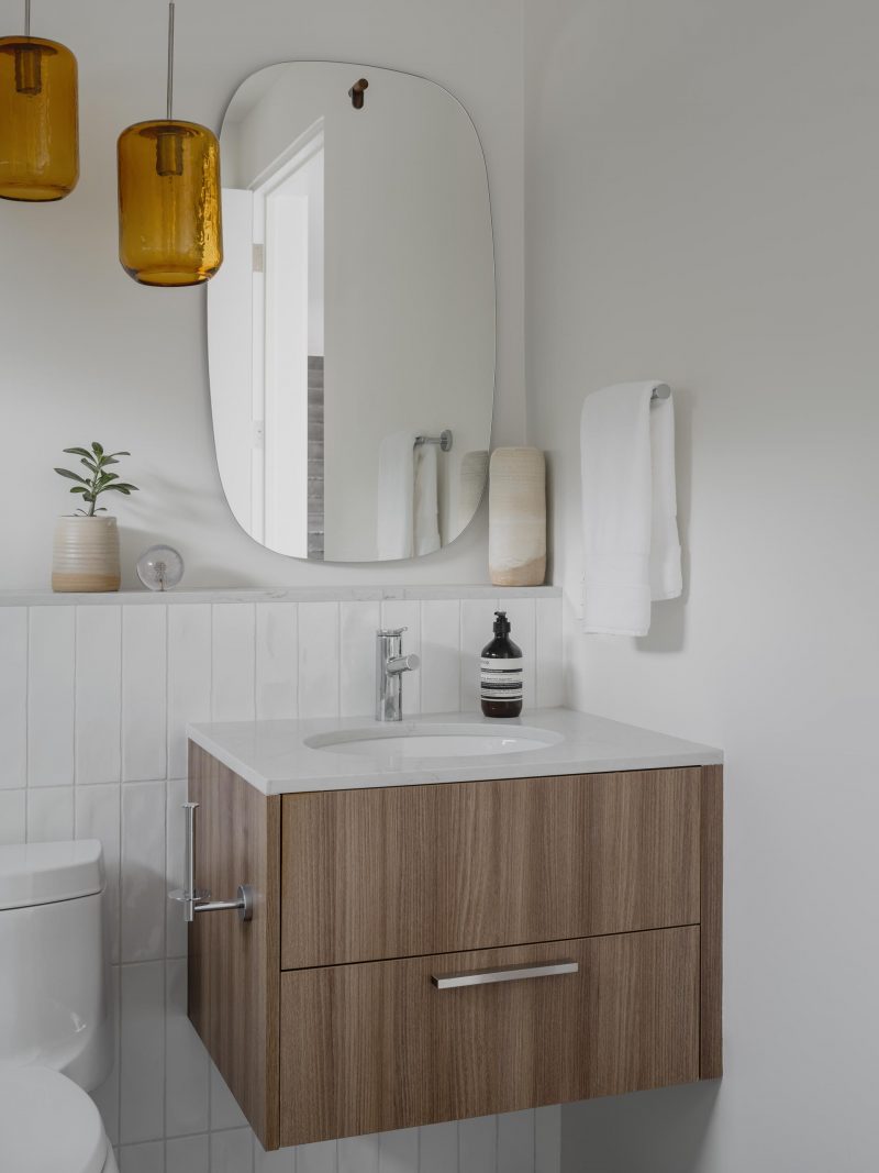 A modern bathroom with white walls and tiles, a floating wood vanity, and a rounded mirror.