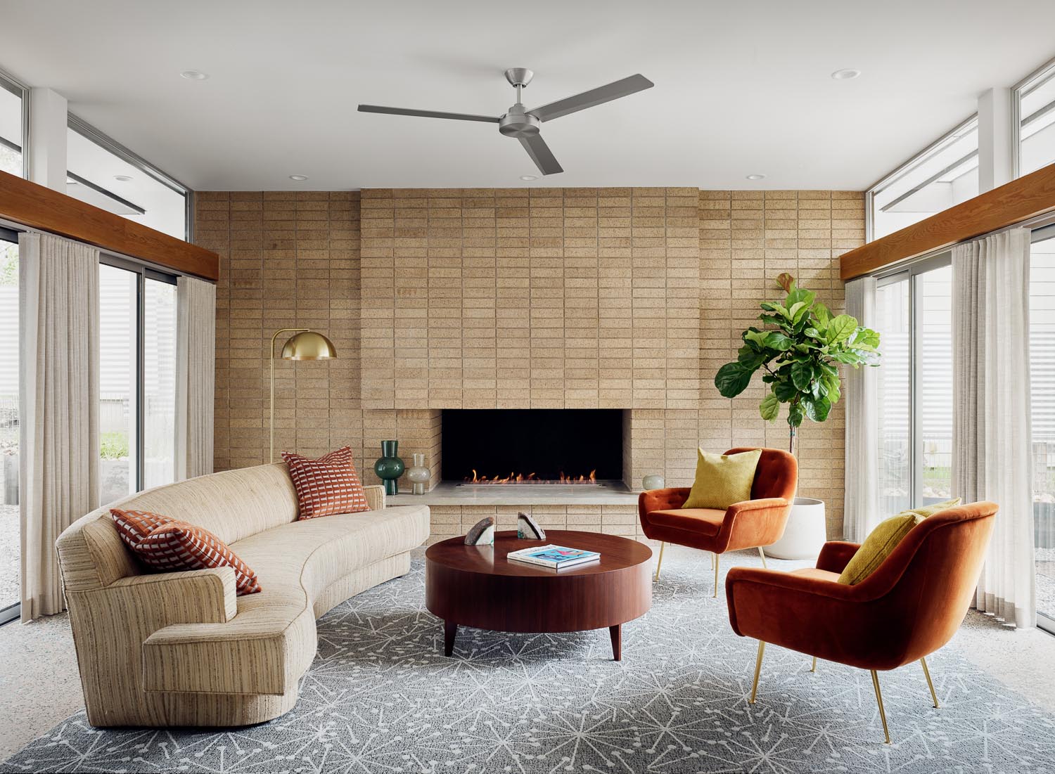 In this mid-century modern inspired living room, there's high ceilings and floor-to-ceiling windows that line the walls, while a brown brick fireplace is the focal point.