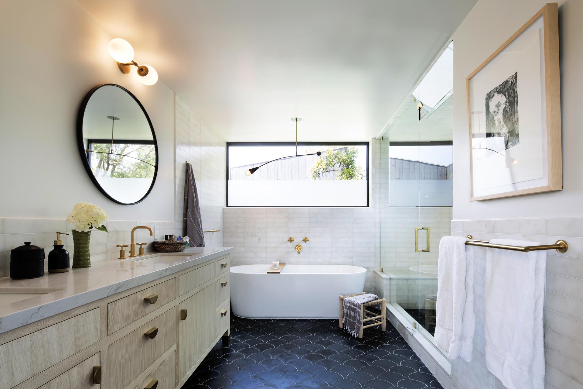 In this modern bathroom there's a double sink vanity with round mirrors, a freestanding bathtub, and a glass enclosed shower.