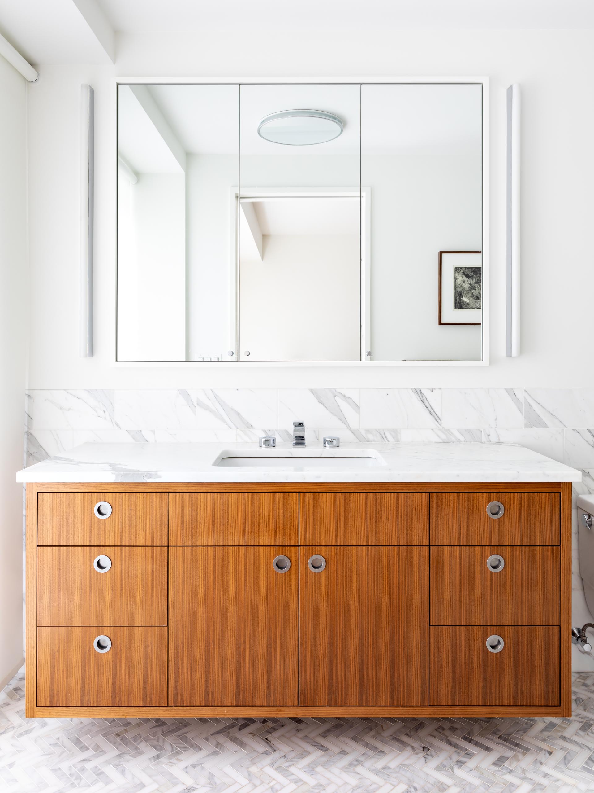 In this mdoern bathroom, there's a floating wood vanity with a tri-panel mirror mounted to the wall, and a marble backsplash that continues through to the shower.