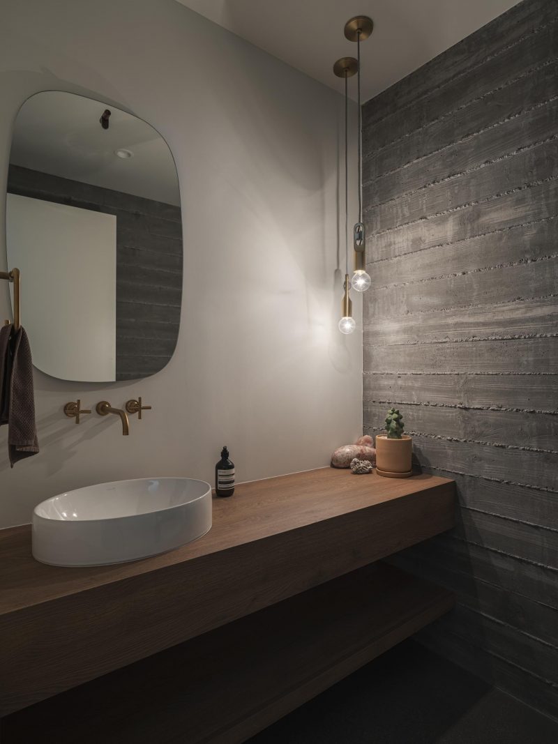 A modern bathroom with board formed concrete wall, a wood vanity, and a rounded mirror.