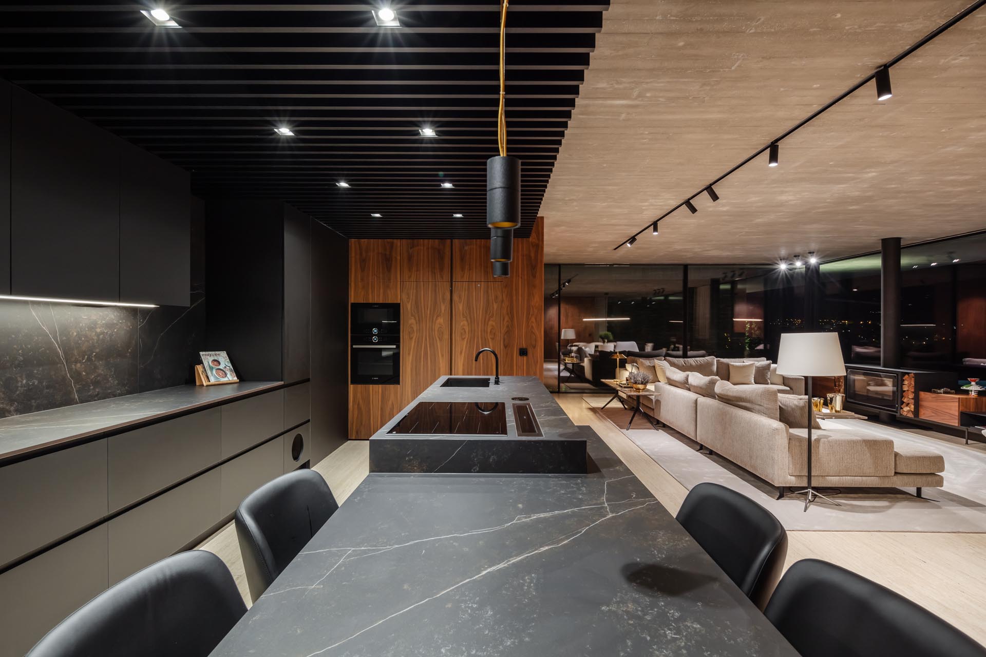 A modern black kitchen includes a large island with dark gray stone, seating for four people, a cooktop, and a undermount sink. 