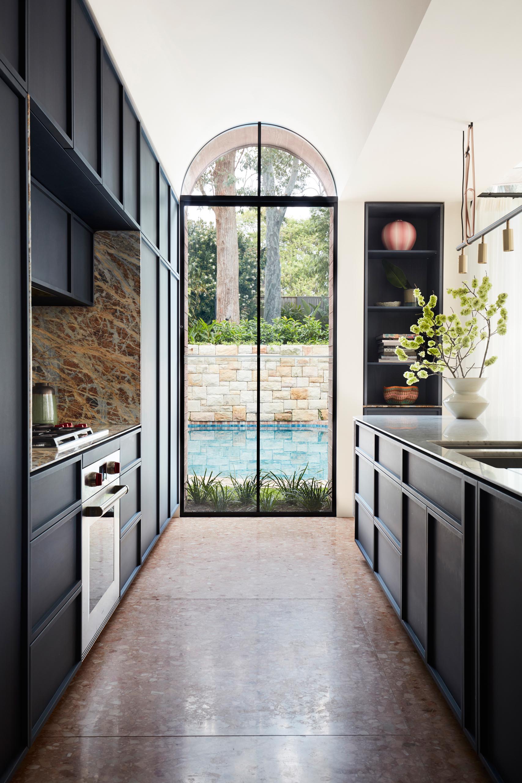 A modern kitchen with dark hardware free cabinets, and a large arched window with views of the swimming pool.