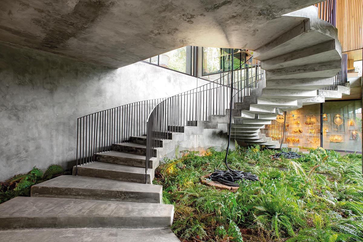 An oversized black-framed glass door, stairs with black railings, wall boxes displaying sculptural items, and gardens, help to create a grand entrance for this modern concrete home.