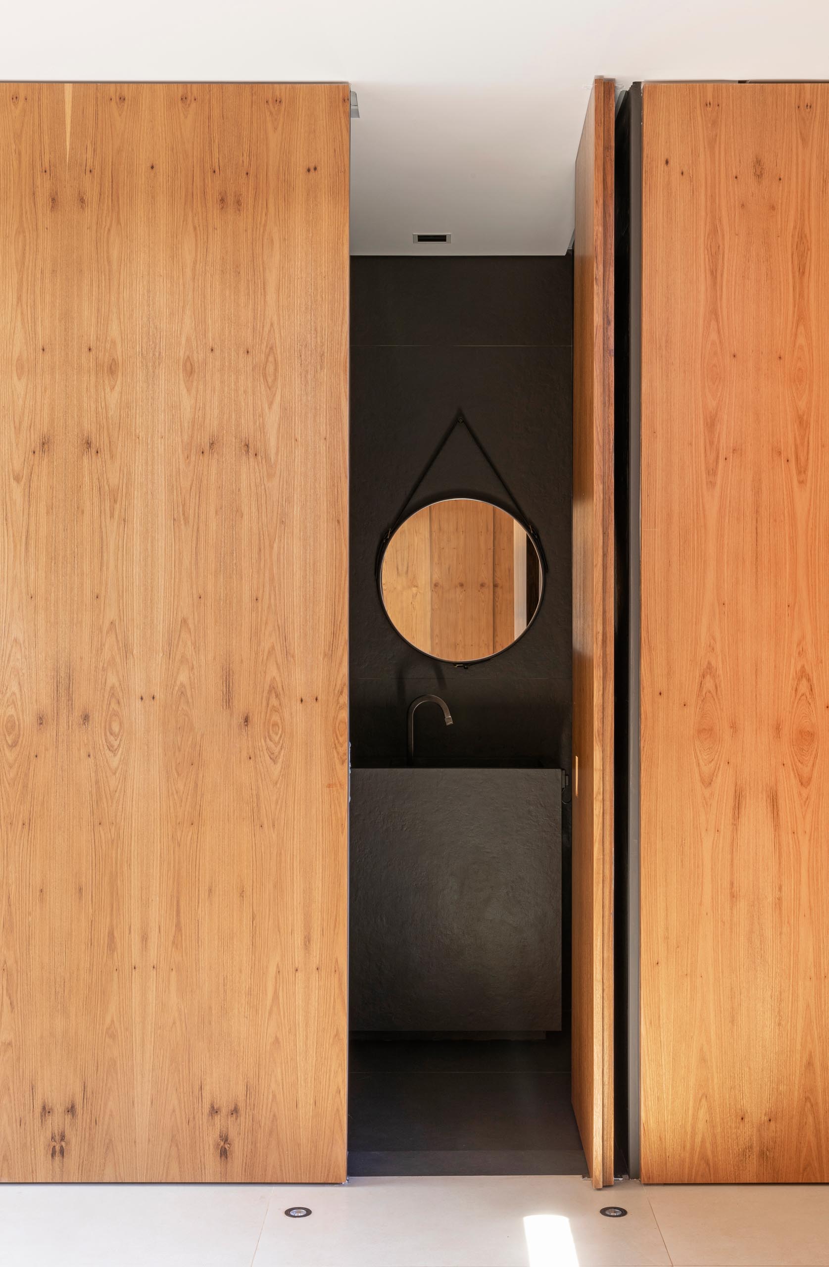 A modern bathroom with a dark color palette is hidden behind a wood door that matches the surrounding wall.