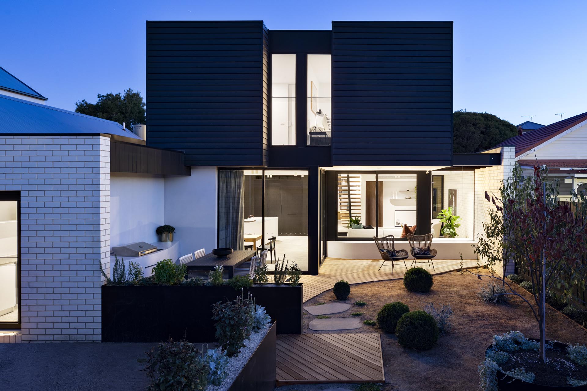 A modern house extension that has a black facade, a landscaped yard with a bbq, outdoor dining area, and patio.