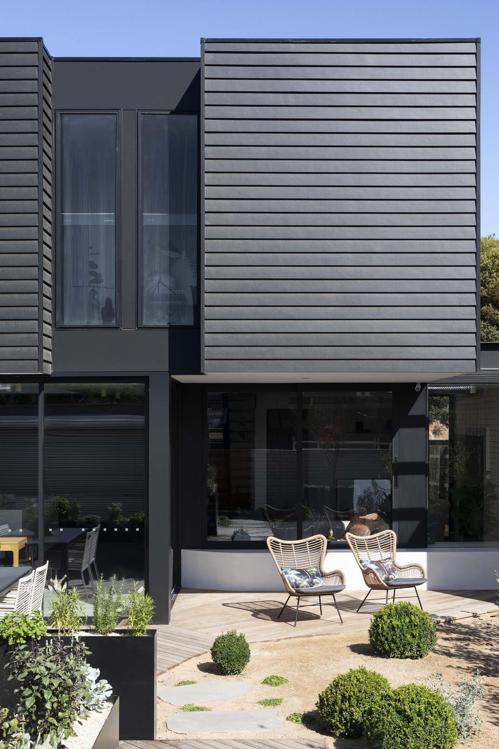 A modern house extension that has a black facade, a landscaped yard with a bbq, outdoor dining area, and patio.