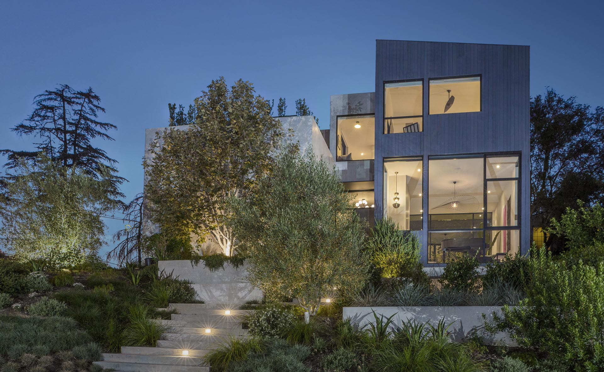 Mature landscaping is separated by concrete stairs that lead up to the house, and at night, lighting creates a guided path.