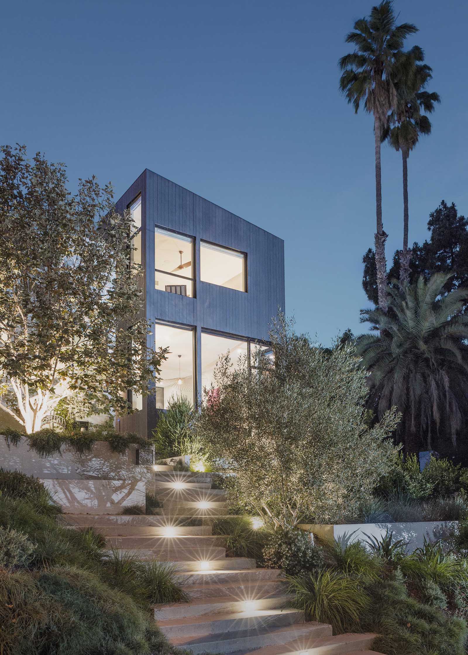 Mature landscaping is separated by concrete stairs that lead up to the house, and at night, lighting creates a guided path.