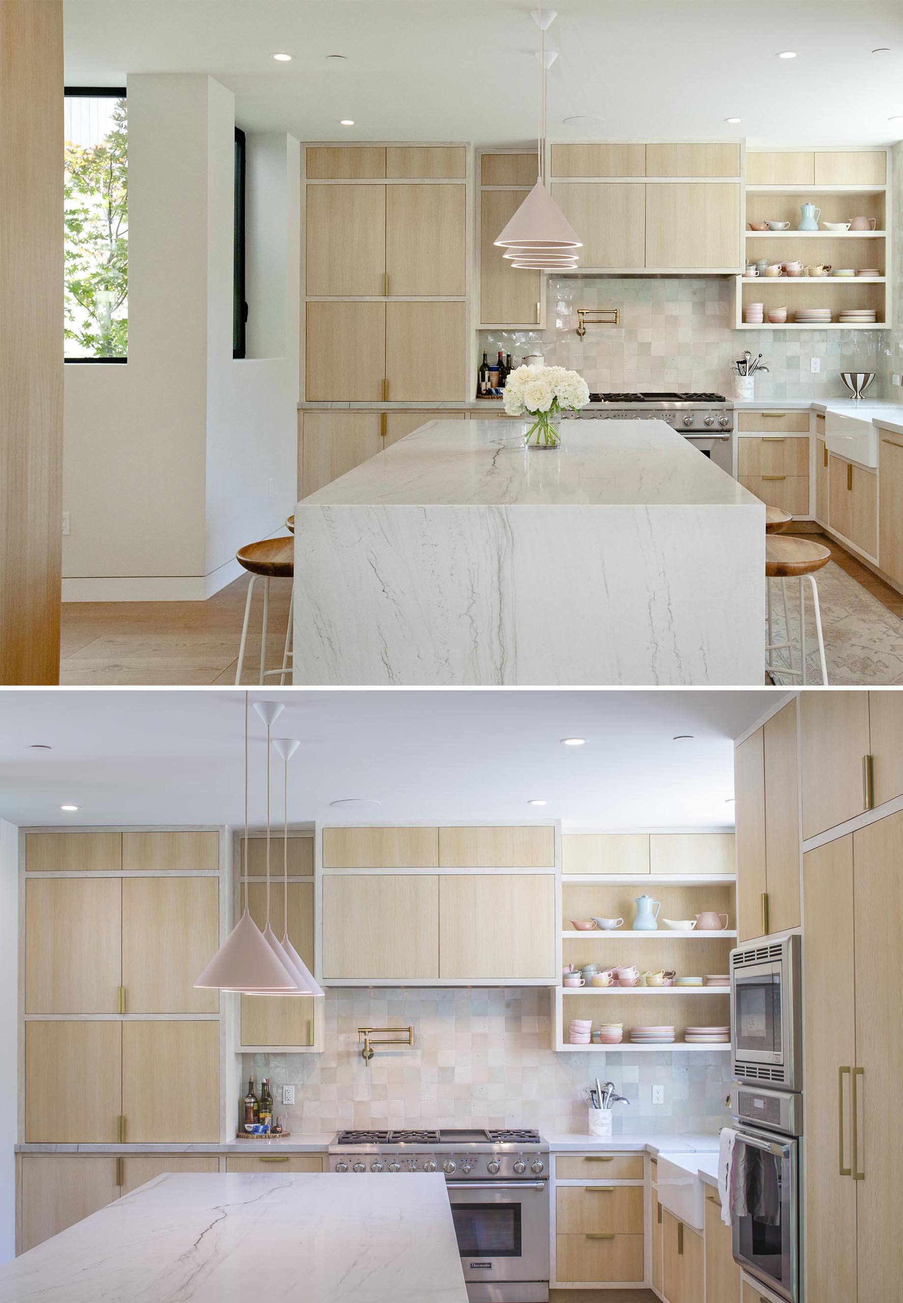 In this modern kitchen, light wood cabinets sit flush within their frames, while an island has a waterfall countertop, and the pastel pink pendant lights complement the displayed dishware and the backsplash.