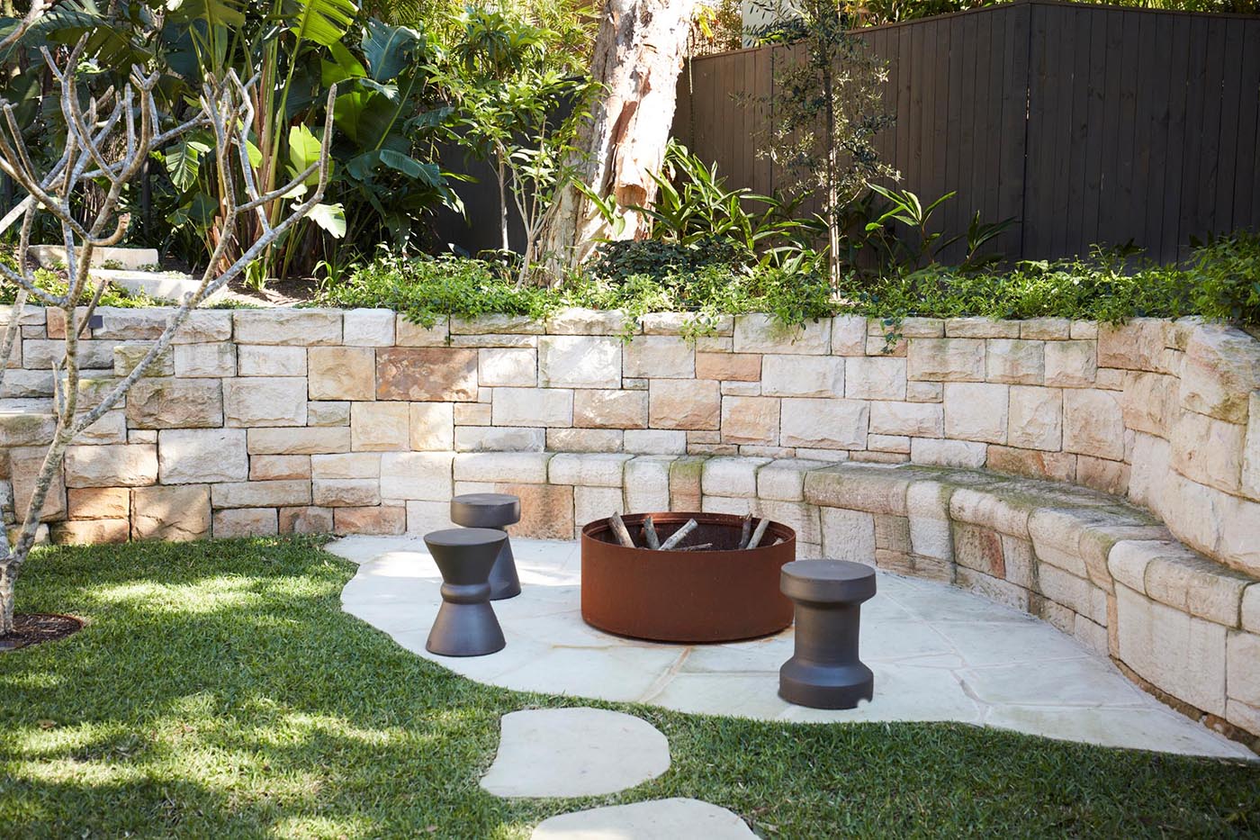 A sandstone wall incorporates seating around a fire pit, and includes steps that lead up to an upper level of the garden.