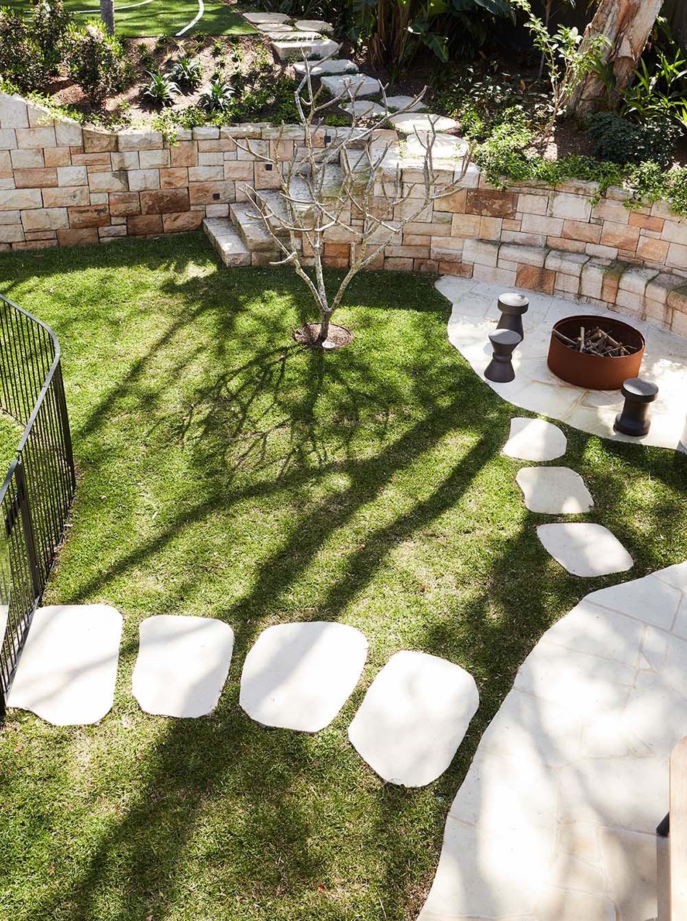 A sandstone wall incorporates seating around a fire pit, and includes steps that lead up to an upper level of the garden.