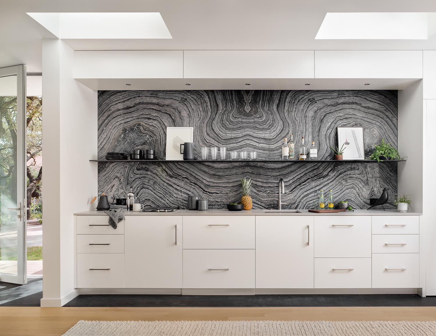 A modern small kitchen with white cabinets and a gray stone backsplash with a minimalist black shelf. Two skylights help to keep the kitchen bright.