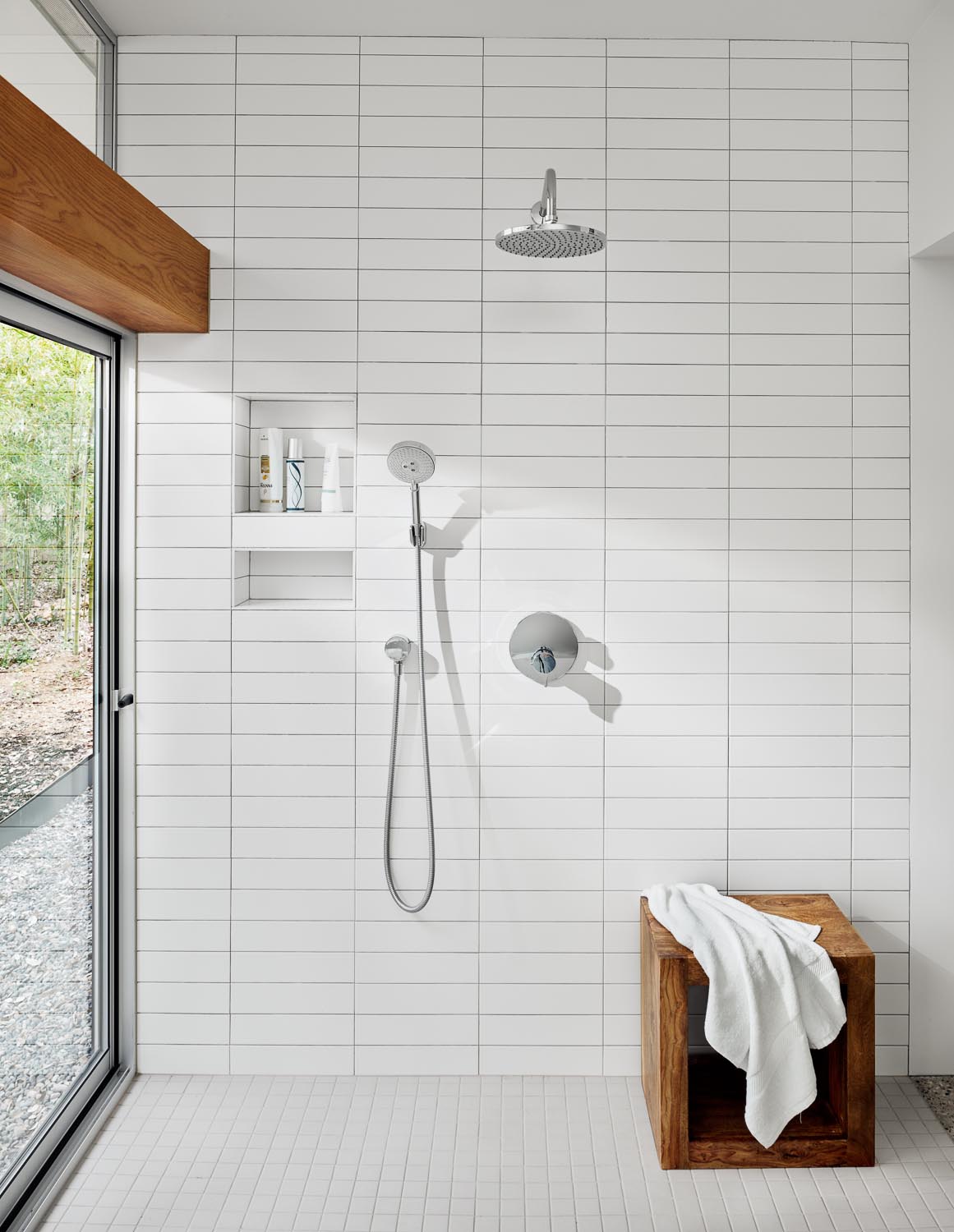 In this modern bathroom, there's a floating wood vanity, a built-in bathtub, and walls lined with white rectangular tiles.