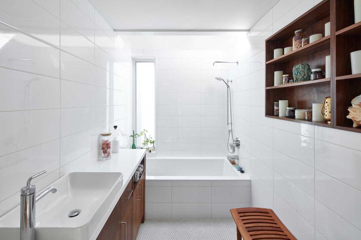 In this modern bathroom, white tiles cover the walls and the front of the built-in bathtub. Wood shelving on the wall complements the wood vanity on the opposite wall. 