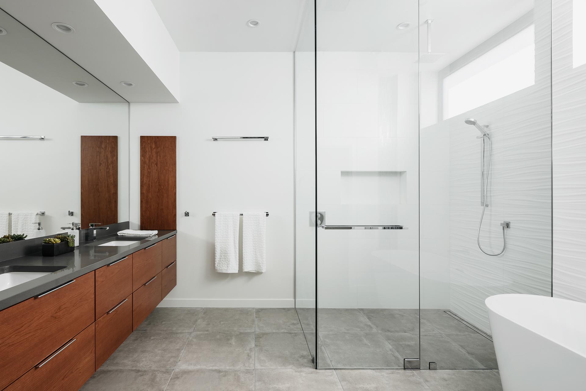 In this modern master bathroom, a floating wood vanity is topped with dark gray quartz counters, a step free shower with frameless glass is positioned in the corner, and an adjacent free-standing tub has a backdrop of a 3d tile wall.