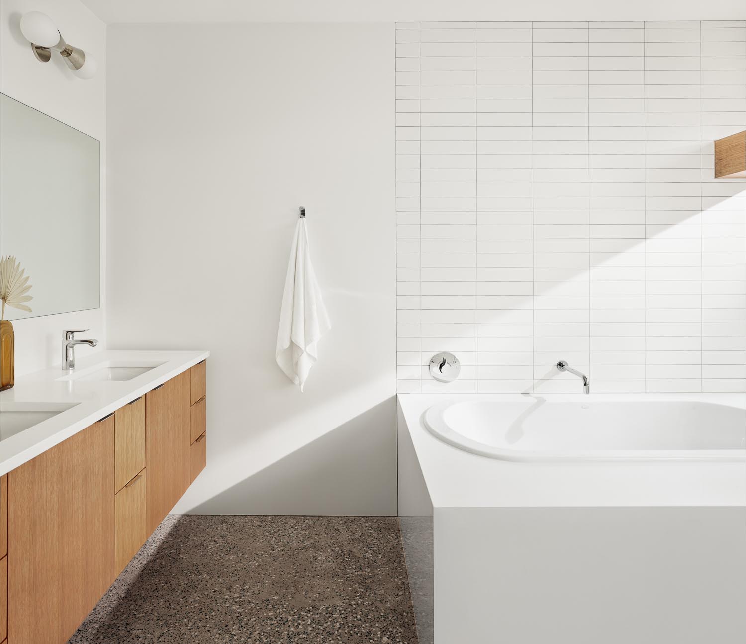 In this modern bathroom, there's a floating wood vanity, a built-in bathtub, and walls lined with white rectangular tiles.