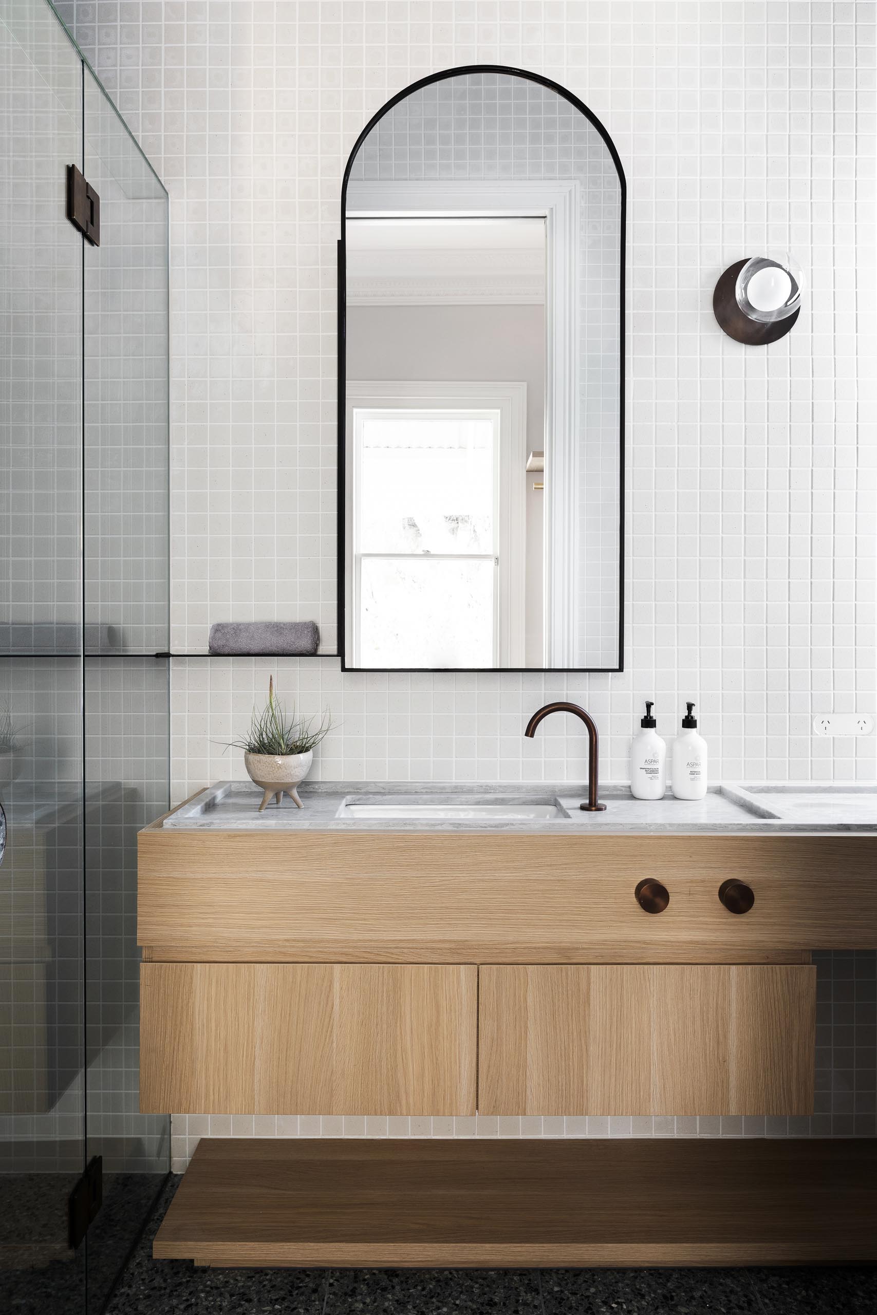 A modern bathroom with a wood vanity, tiled wall, and an arched mirror.