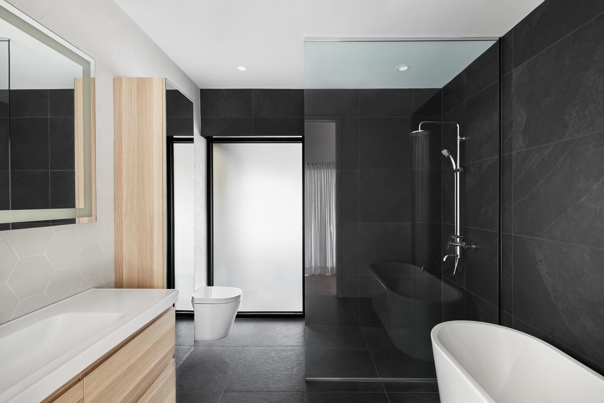 In this modern bathroom, large format black tiles cover two of the walls, while there's a light wood vanity, and a wall of white hexagonal tiles.