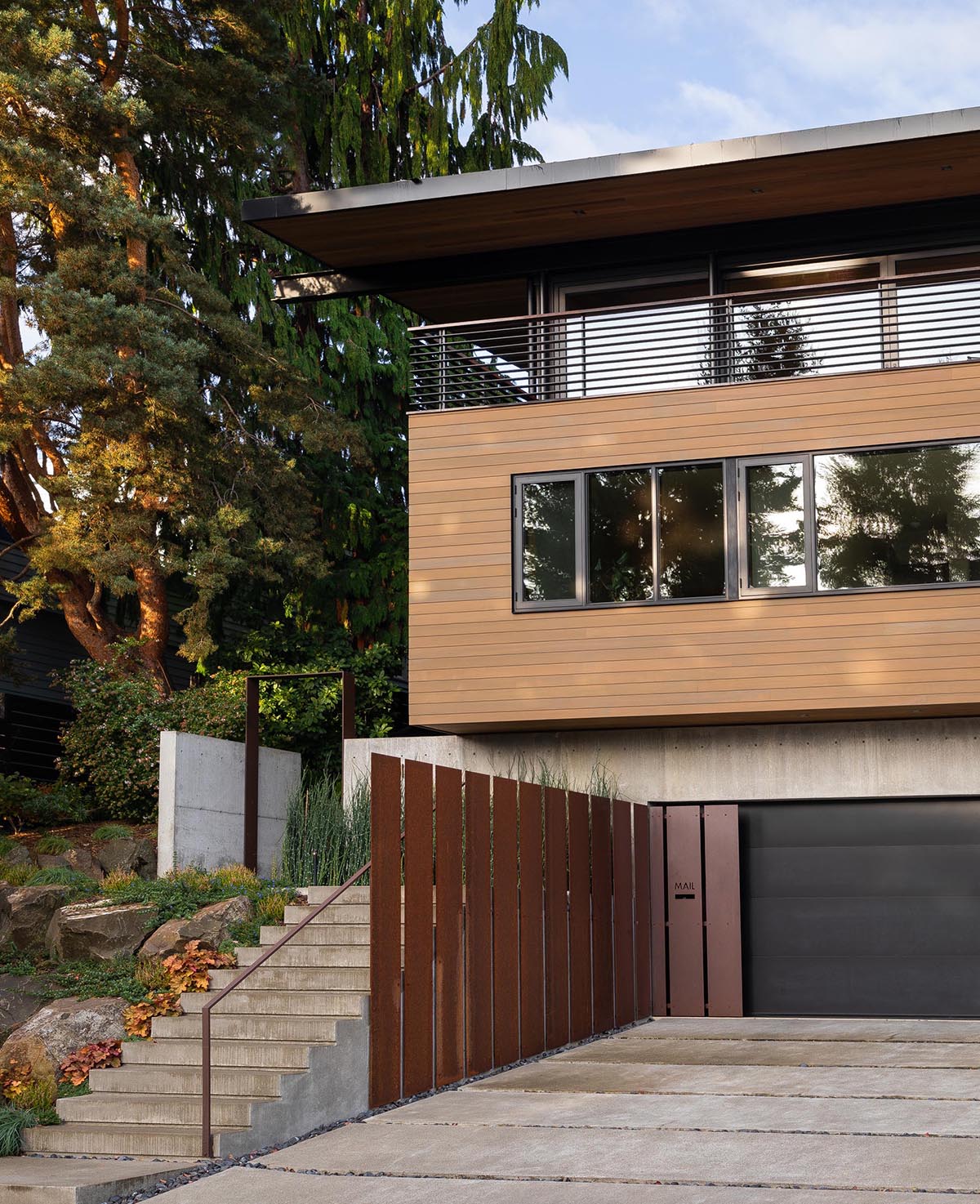 The facade of this modern house features a concrete, a black garage door, steel accents, wood siding, and steps that lead to the front door.