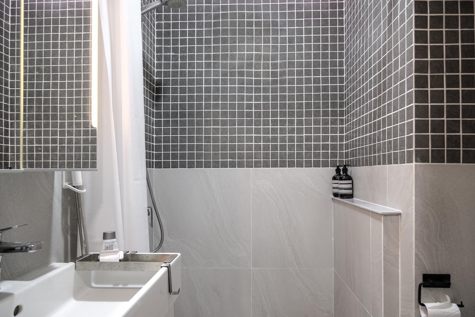 In this small bathroom, large tiles cover the lower half of the walls, while the upper half is covered in square gray tiles.