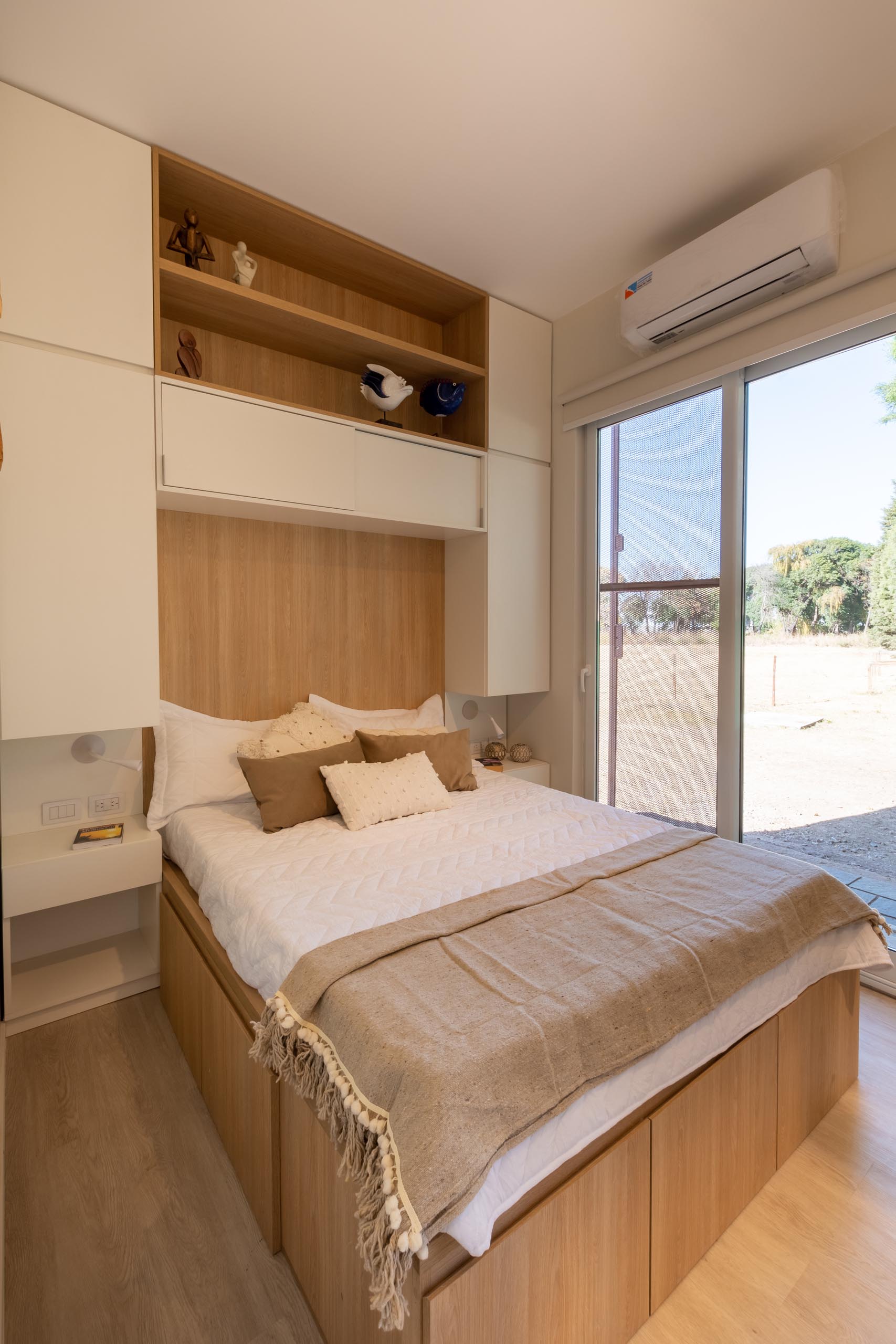 In this tiny home, there's a bedroom with a wood accent and shelving surrounded by white cabinetry. Opposite the bed is a closet with a mirrored front.
