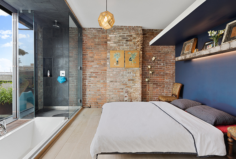 This primary bedroom has a deep blue accent wall behind the bed, while the bathroom is completely open to the bedroom, and includes a sunken bathtub with a rolling wood cover and a glass enclosed shower.