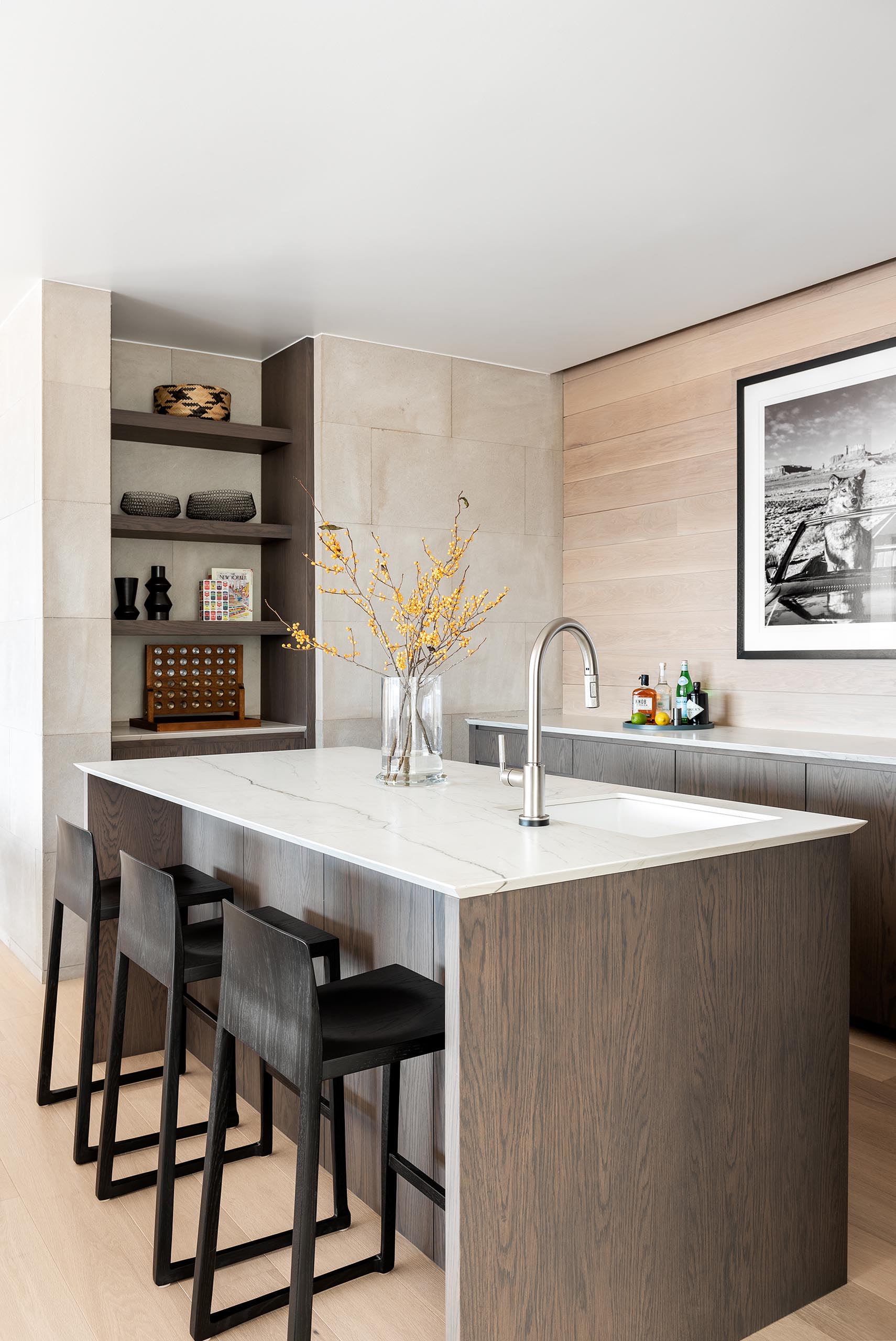 A modern bar with an island, wood cabinets, open shelving, and light colored countertops.