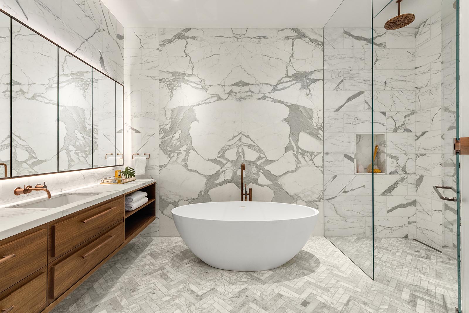 This modern master bathroom includes gray and white book-matched marble slabs on the walls, a floating wood vanity, a freestanding Badeloft bathtub, and a glass enclosed shower.