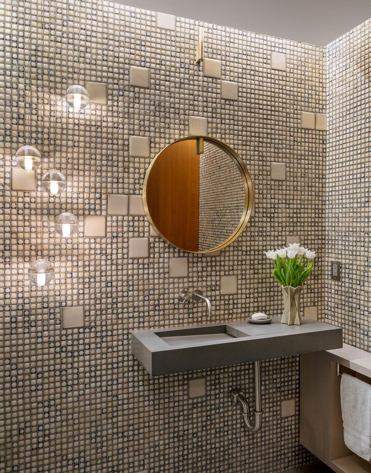 In this powder room, there's a combination of small and large tiles covering the walls, while a round mirror hangs above a concrete vanity.