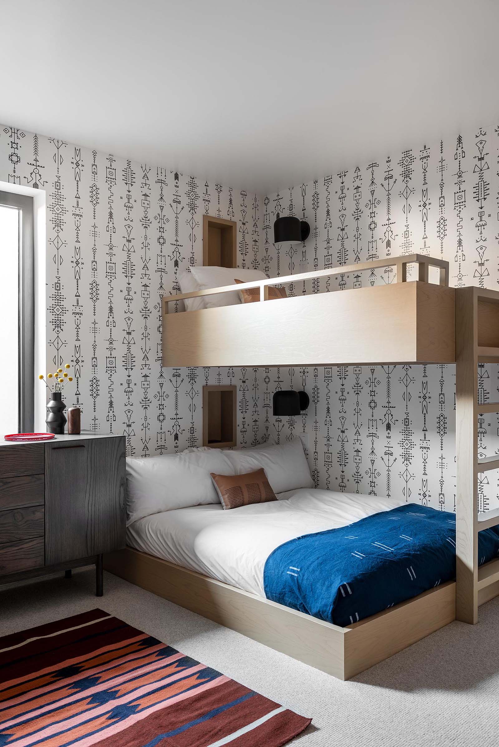 In this modern bedroom, a graphic wallpaper wraps around the room, while bunk beds are designed to sleep three people, and each bed has a small niche and wall mounted matte black light.