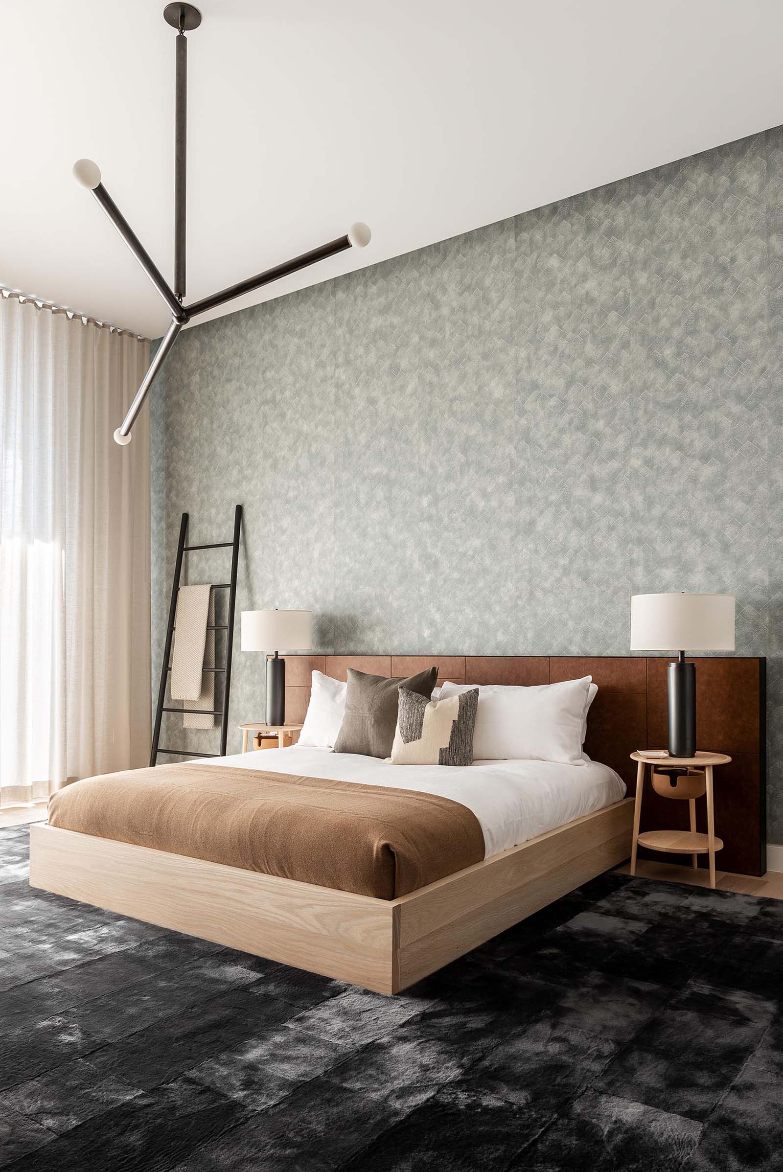 In this modern bedroom, there's a muted gray wallpaper that covers the entire wall, creating a backdrop for the bed, while minimalist pendant light hangs from the ceiling, and a matte black ladder provides a place to hang blankets.