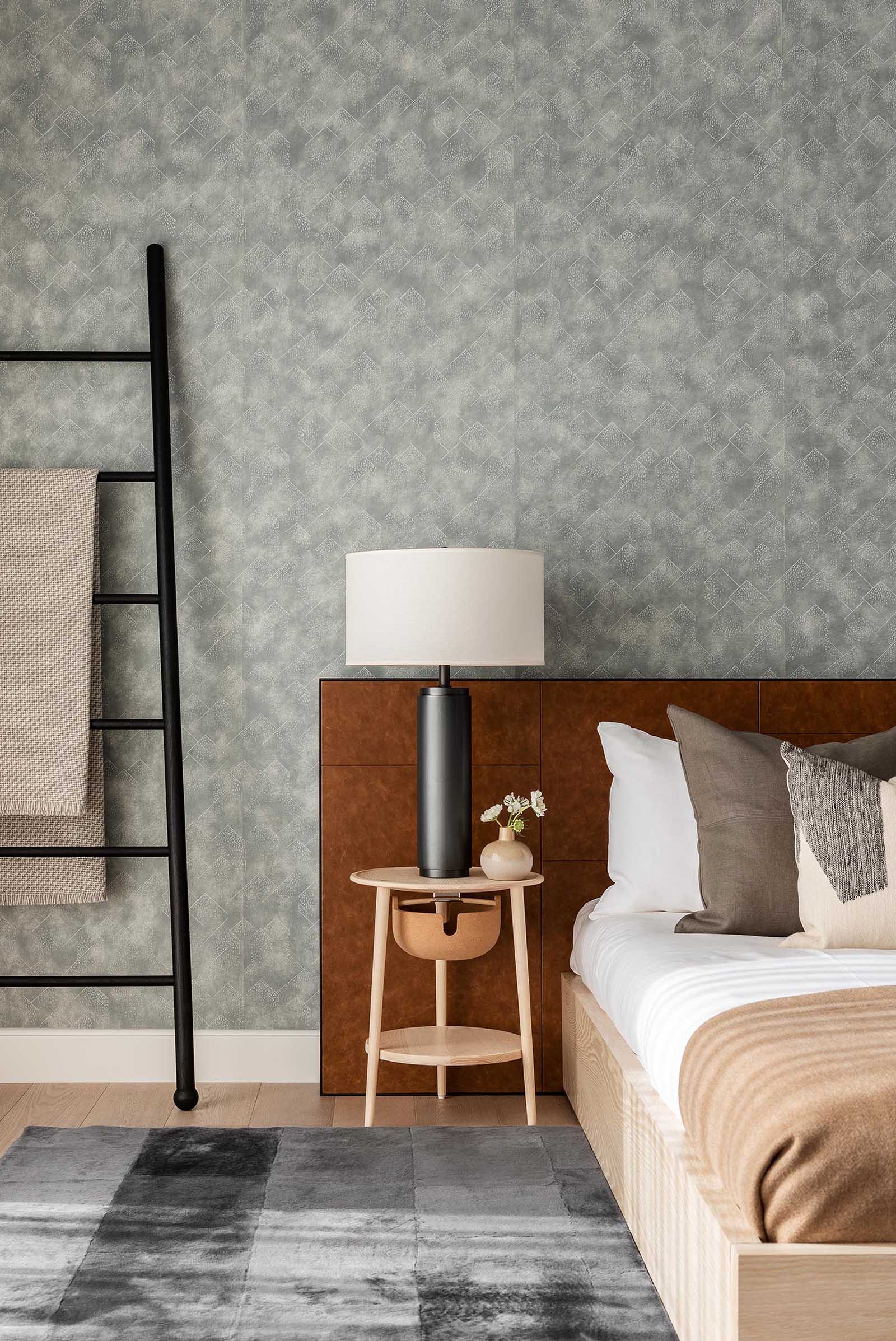 In this modern bedroom, there's a muted gray wallpaper that covers the entire wall, creating a backdrop for the bed, while minimalist pendant light hangs from the ceiling, and a matte black ladder provides a place to hang blankets.