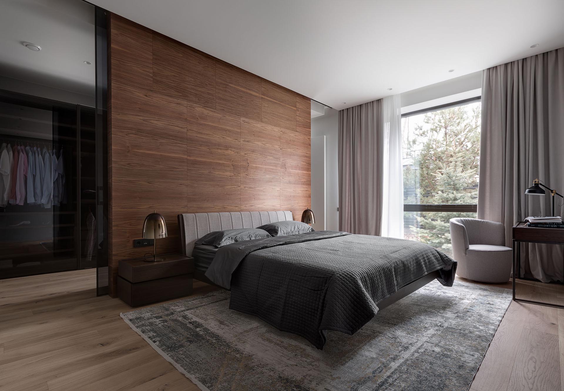 In this modern primary bedroom suite, there's a wood accent wall that separates the bedroom from the walk-through closet, that also includes black glass doors.