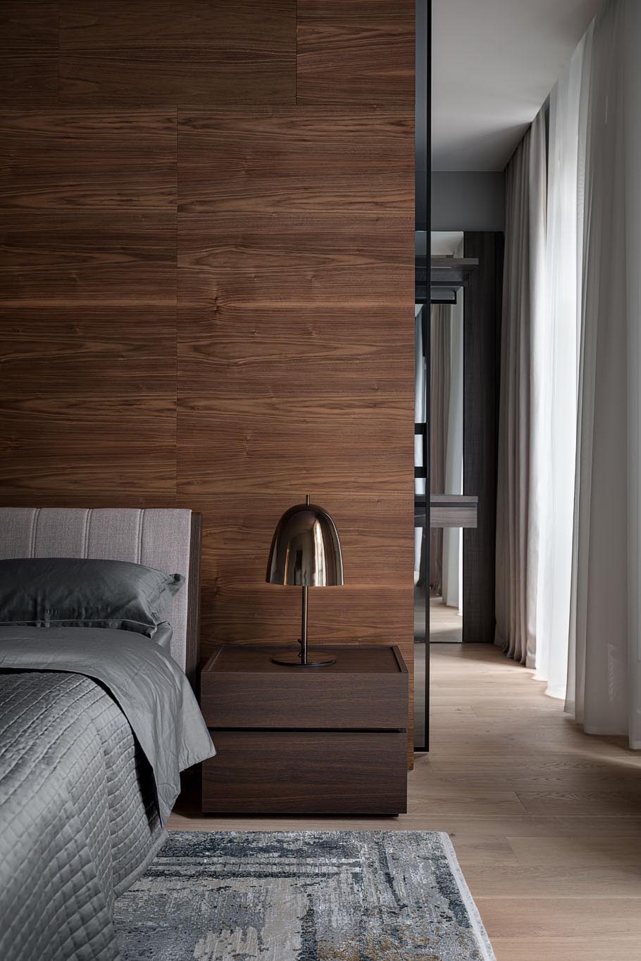 In this modern primary bedroom suite, there's a wood accent wall that separates the bedroom from the walk-through closet, that also includes black glass doors.