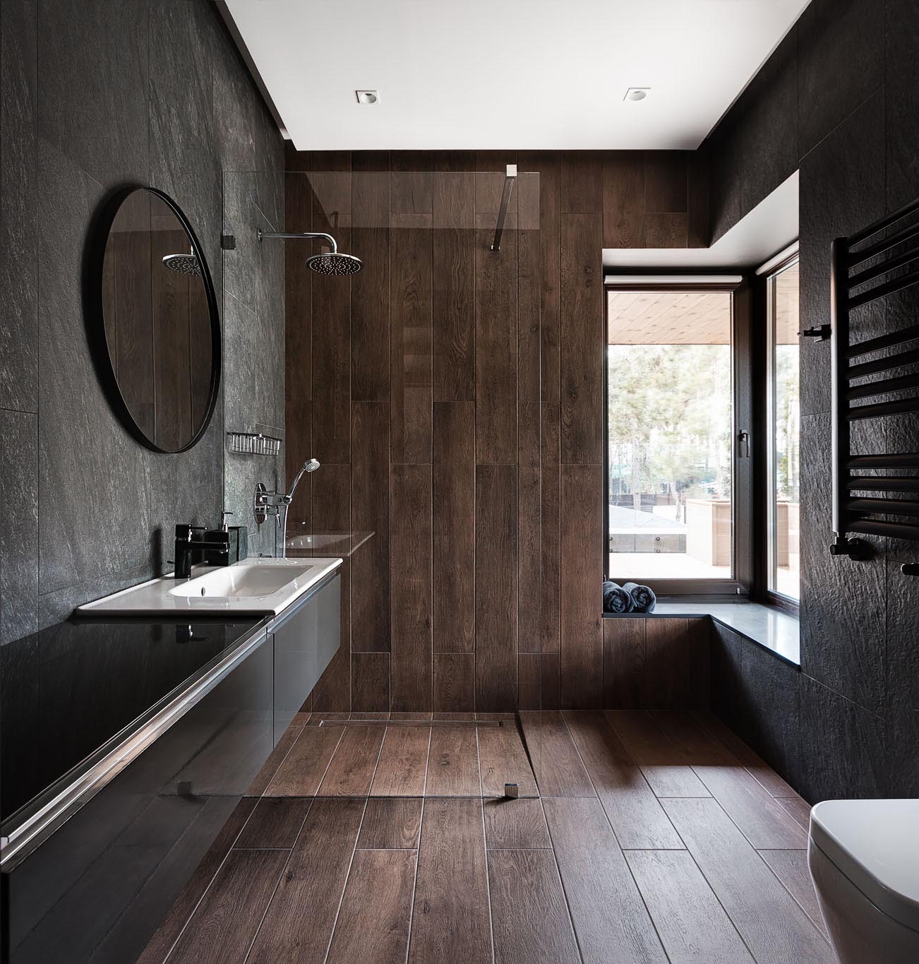 This modern bathroom has a dark material palette with gray walls, wood tiles that travel from the floor to the wall, and a floating black vanity.