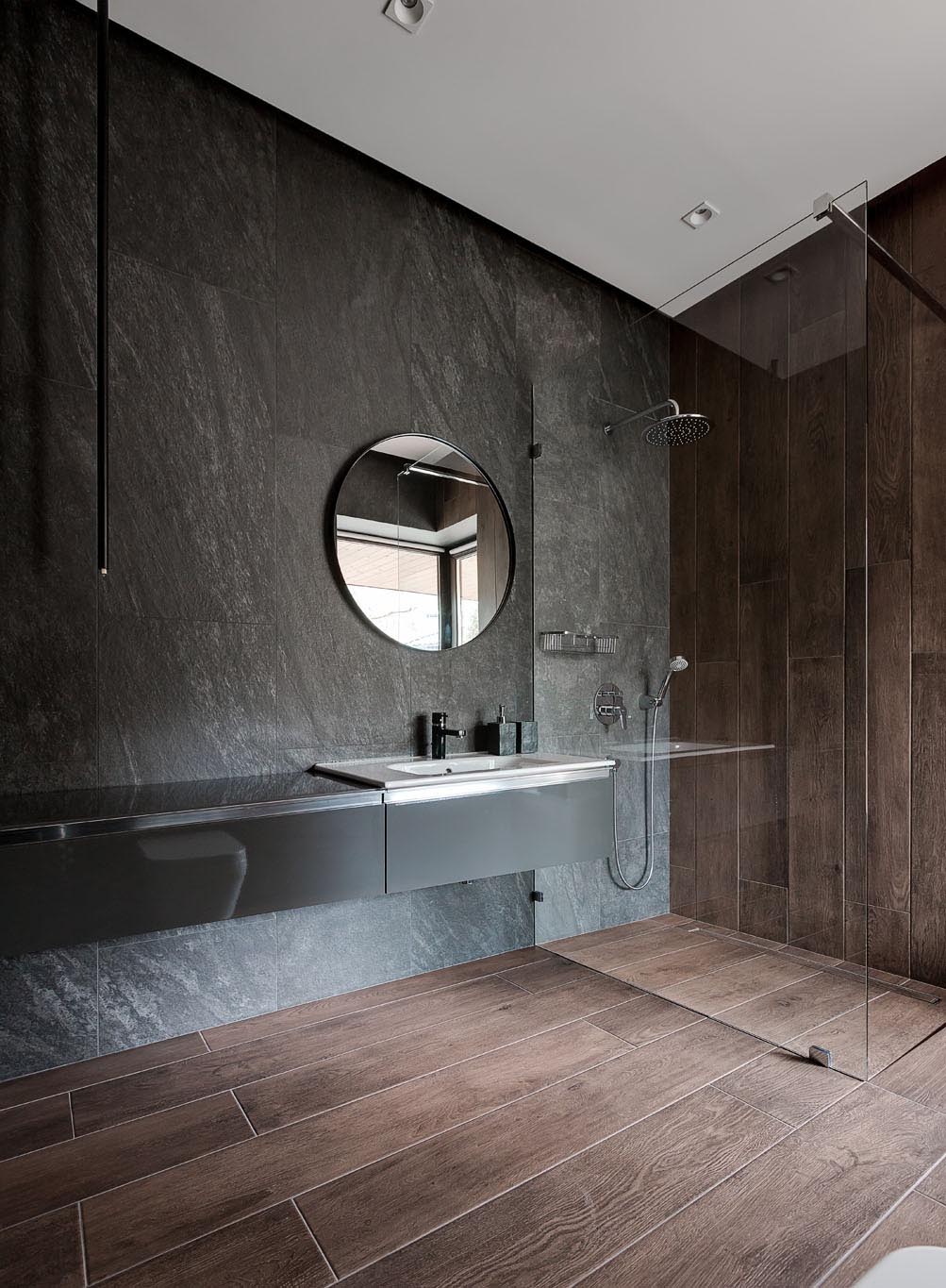 This modern bathroom has a dark material palette with gray walls, wood tiles that travel from the floor to the wall, and a floating black vanity.