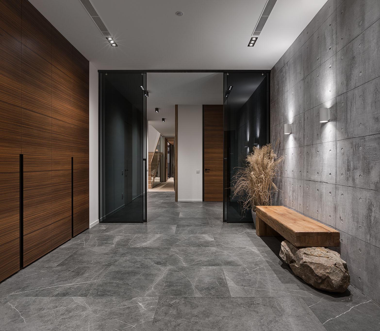 This modern entryway has an exposed concrete wall highlighted by two wall scones, as well as a dark wood wall, and features a wood bench that rests on a boulder.