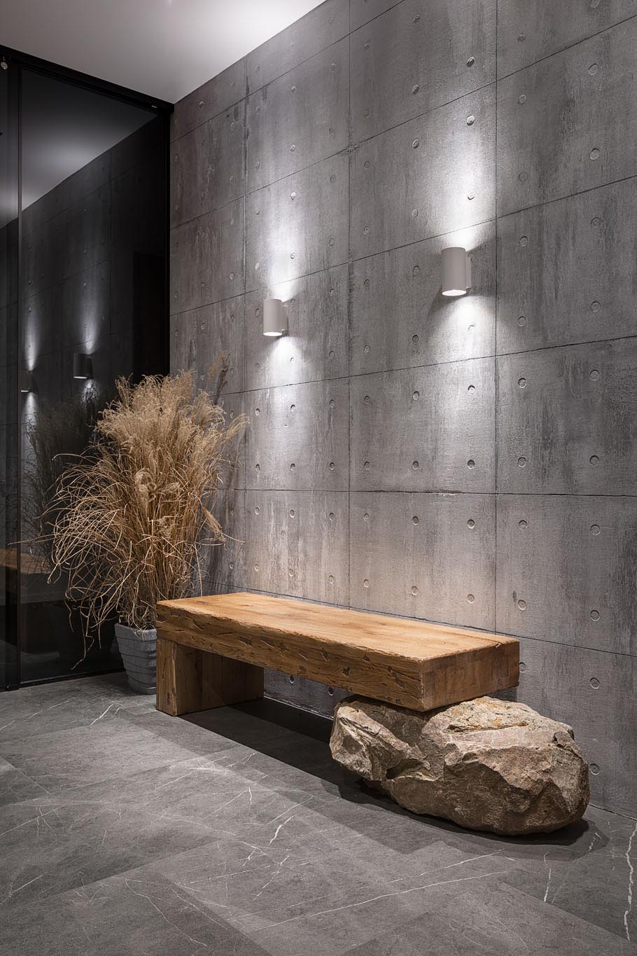 This modern entryway has an exposed concrete wall highlighted by two wall scones, and features a wood bench that rests on a boulder.