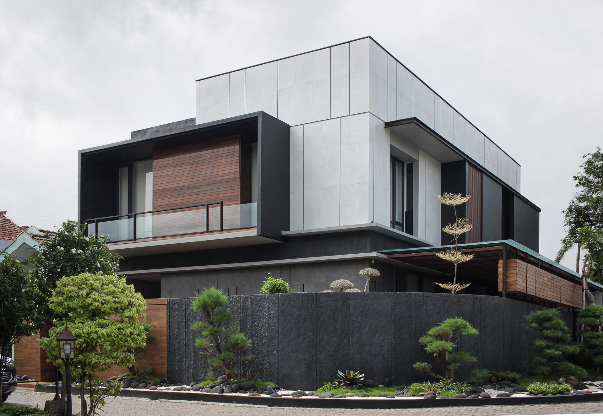 This Zen garden creates modern  curb appeal and has a dark matte black and wood fence as a backdrop. It also includes ornamental trees, white pebbles, gray stones, and small sections of grass.