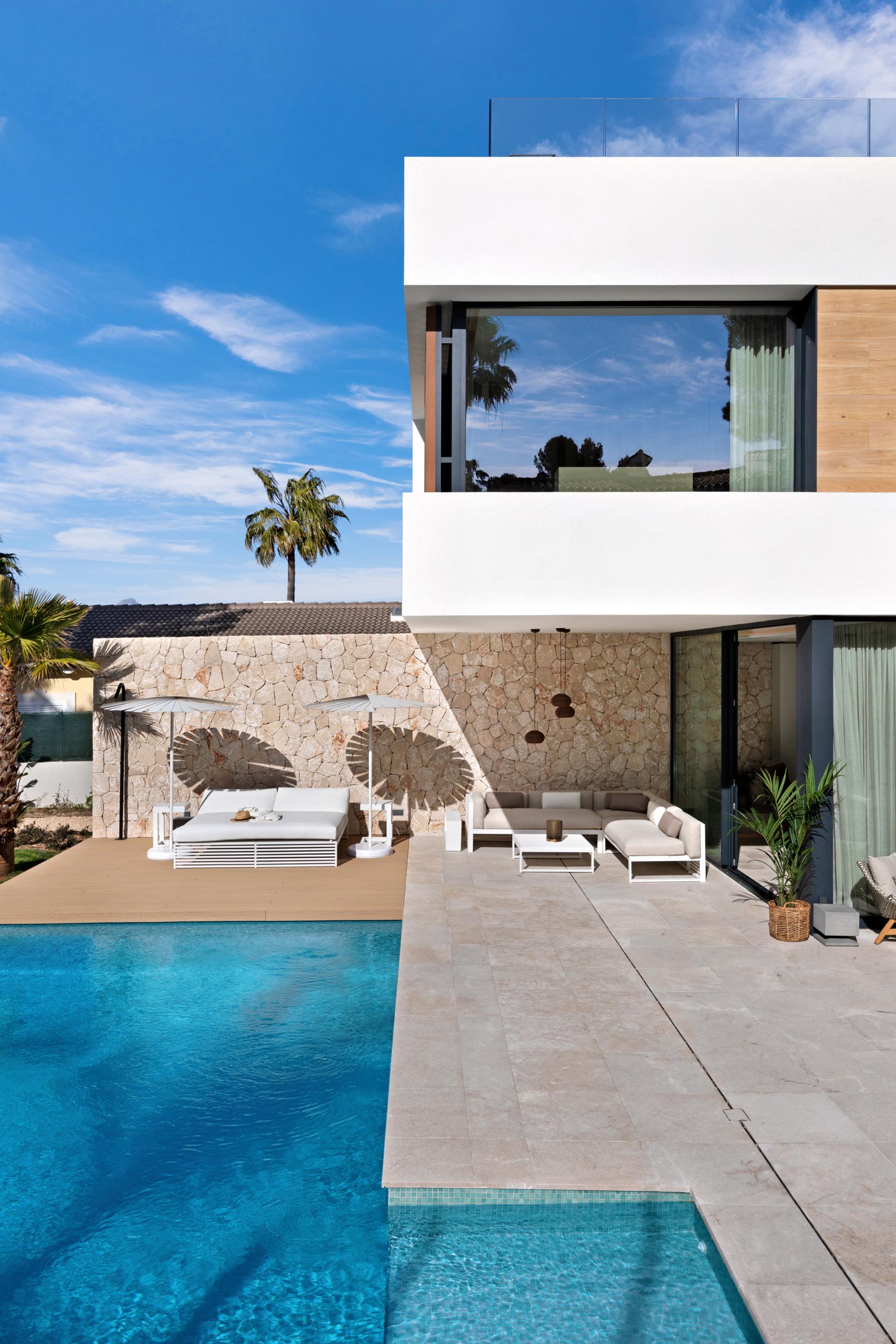 A modern home with a swimming pool and deck, a stone wall, and partially covered patio designed for outdoor entertaining.