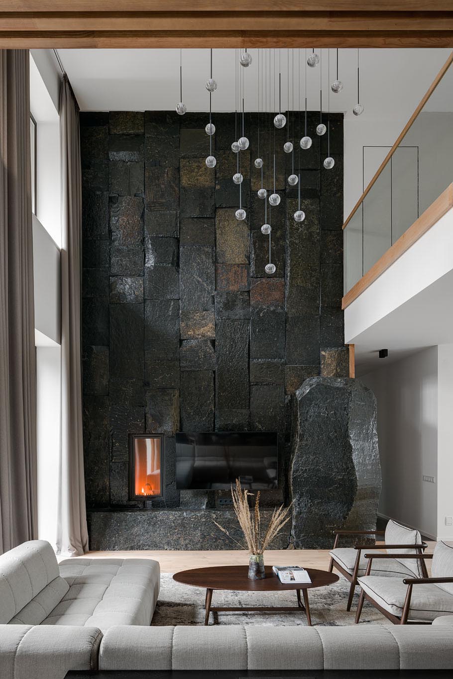 In this modern living room, there's a double height ceiling with a sculptural light installation, and a large, dark, and monumental natural stone boulder wall that's used as a backdrop for the fireplace and television.