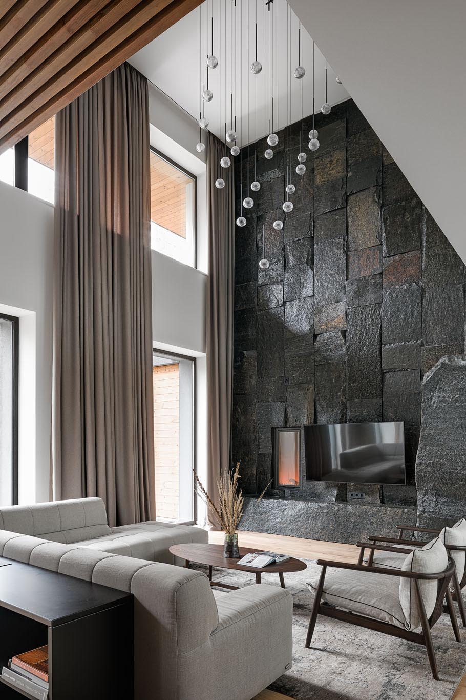 In this modern living room, there's a double height ceiling with a sculptural light installation, and a large, dark, and monumental natural stone boulder wall that's used as a backdrop for the fireplace and television.