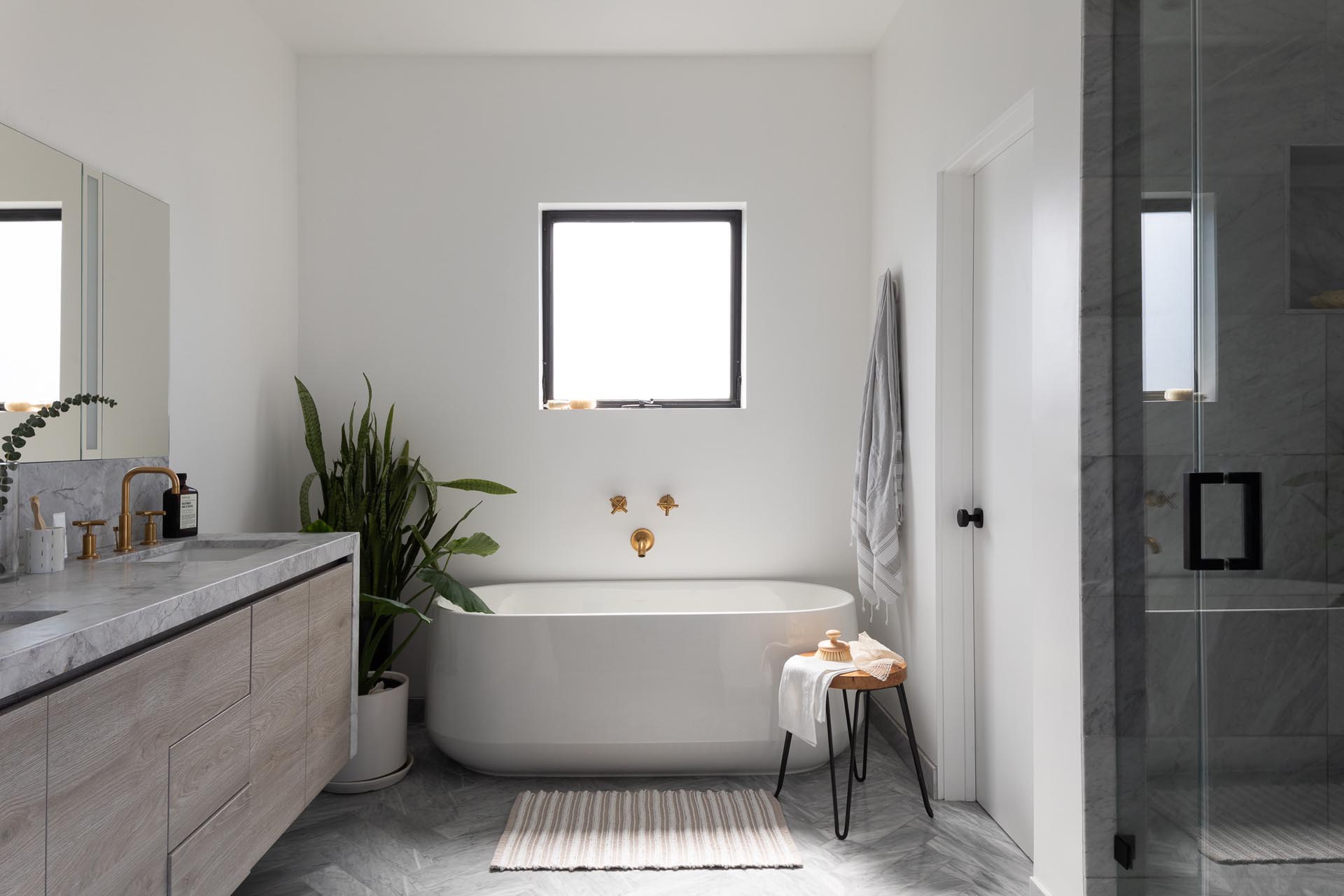 In this modern en-suite bathroom, there's a freestanding white bathtub, a walk-in shower, and a wood vanity with a waterfall countertop.