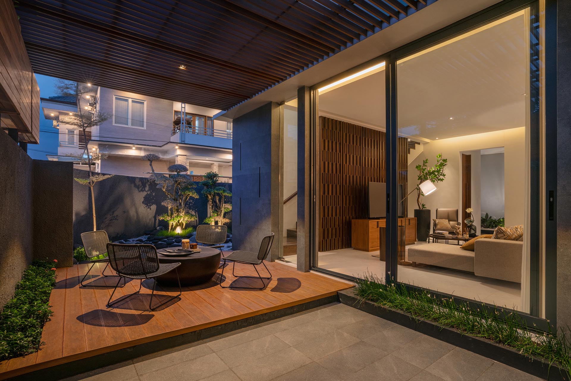 A partially covered patio has a wood deck that's furnished with a round table and outdoor chairs. Lining the interior wall of the fence is similar landscaping to the outside of the house.