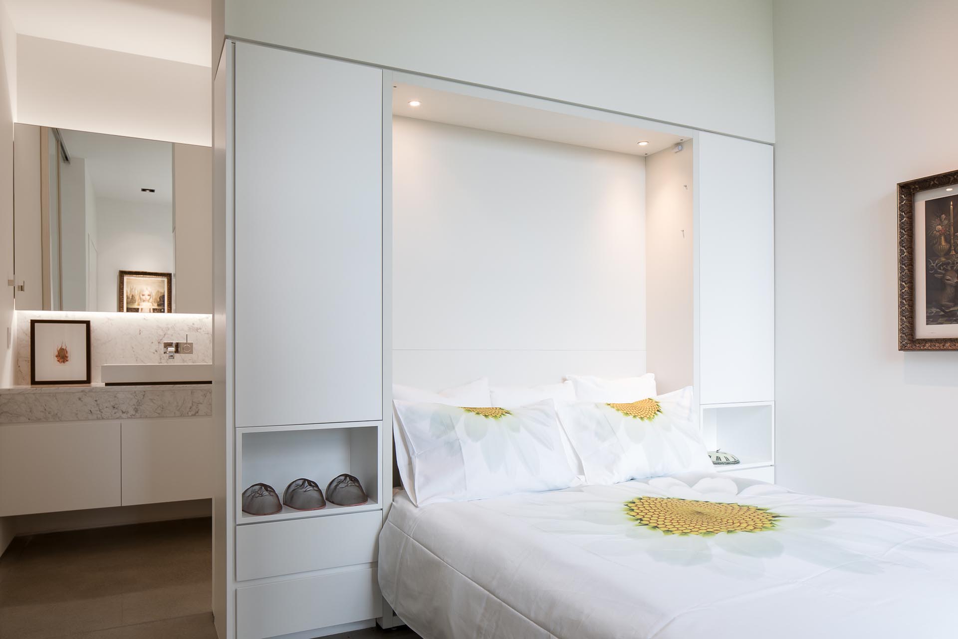 In this modern white bedroom, custom cabinetry surrounds the headboard of the bed, and separates the sleeping area from the en-suite bathroom.